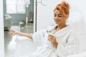 waist-up-portrait-view-charming-woman-white-bathrobe-sitting-armchair-receiving-iv-infusion-she-is-holding-glass-lemon-beverage-smiling